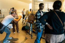 In a museum gallery, students exhibit projects on easels. Two students speak with adults about their work.