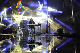 A performer is sitting behind a keyboard on stage with beams of blue and white lights and a rainbow flag flying on a pole.