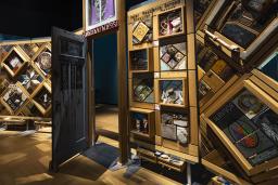 A large, curved freestanding wooden wall on which is mounted numerous small objects arranged in diamond and rectangular patterns. There is an open door in the centre of the wall.