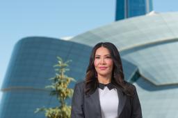 A woman with long dark hair, wearing a suit jacket, stands with her hands clasped in front of her. Behind her is a large, unusual building with a rounded glass façade and a tower.