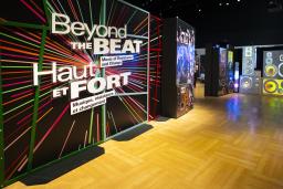 The entrance to a museum exhibit with a graphic panel includes colourful rays and text that reads “Beyond the Beat: Music of Resistance and Change” in English and French. A projected image of a pile of various speakers fills the adjacent wall.