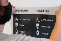 A man holds up a sign that says "Washroom" as it is mounted to a wall. Underneath the title, the words "accessible," "toilet" and "urinal" are accompanied by descriptive icons.