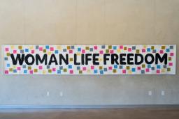 A white banner hangs on a wall, bearing the words "Woman, Life, Freedom" in black. Small coloured squares with text are attached to the banner around the text.