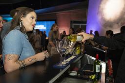 A woman waits for a drink at a bar in a museum space, while a bartender pours glasses of wine.