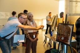 Students in a museum gallery display their work on easels. The focus is on one student talking with an adult who is smiling and taking a photograph with their phone of the student’s work. The artwork shows four people in a hallway with four doors, each with a sign on it.