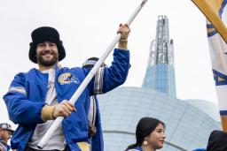 A smiling man in a crowd of people holding a large yellow flag with a blue and white, “W” in the centre. The man is wearing a blue Winnipeg Blue Bombers jacket as he stands in front of the Canadian Museum for Human Rights.