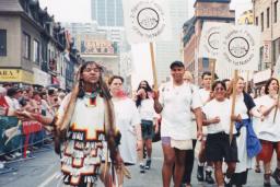 A group of marchers carrying signs that read “2-Spirited People of the 1st Nations” are walking down the centre of a city street while onlookers watch behind barriers on both sides.