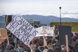 A crowd of people facing away from the camera. Some in the crowd are holding signs above their heads that say: “I can’t breath #BLM”, “Canada is not immune to racism”, and “Justice for all: end violent, subtle and systemic racism now!!!”