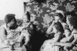 A black and white image of women and young children sitting on couches and chairs in a small living room.