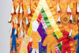 A section of an orange jingle dress is shown. Jingles, or small silver cone-shaped pendants, are visible at the top of the image. Under them, seven human figures are shown, all in various colours and sizes. One of the figures is wearing an orange shirt. These human figures are holding hands inside a multicoloured tipi.