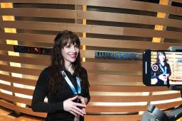 A smiling woman with light skin is filmed on a cell phone. She is wearing a lanyard with the words “Be inspired” and standing in front of an exhibit element made of horizontal strips of wood, some decorated with Indigenous art.
