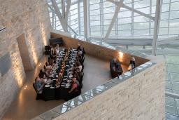 An elegant long table with people dining in an architecturally striking space, featuring tall glass windows and a high ceiling supported by metal beams.