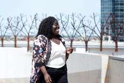 A smiling Black woman stands in front of a row of trees. She is looking to her left.