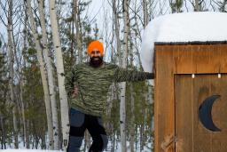 A Sikh-Canadian man, smiling, wearing a green sweater, blue and brown pants, boots, and an orange turban, leaning against a cabin with a crescent moon.