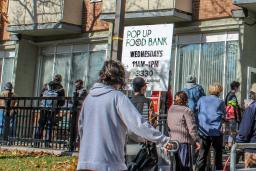 A line-up of people wait outside a building. There are signs indicating it is a Food Bank. A Canadian flag flies above them.