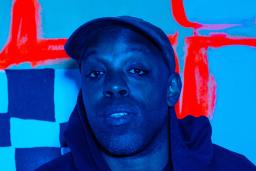 A blue-toned image of a black man wearing a cap and dark hoodie in front of a checkered backdrop with thick red lines.