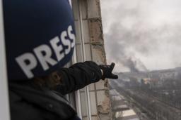 A person wearing a dark blue helmet marked with the word “PRESS” points from a balcony window high above a road, towards a pillar of smoke following an explosion.