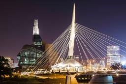 A nighttime shot of an unusual building with a glowing tower, standing beside an illuminated modern bridge, which is reflected in the river water below.