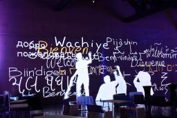 Still image from an animation showing human figures writing “welcome” in multiple languages in white and yellow on a purple wall.