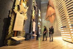 Five museum visitors look at towering exhibits in the “Indigenous Perspectives” gallery at the Canadian Museum for Human Rights. The nearest is made of wood and features trees, animals and a plaque. Behind the visitors, a rounded theatre built of bent wooden slats is visible.