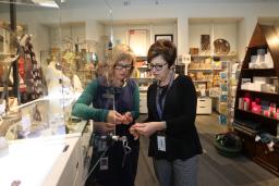 Two women look at a piece of jewellery. They are standing in a store full of colourful merchandise.