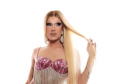A glamourous drag queen with long blonde hair and dramatic makeup is wearing a bejeweled red bikini top with long silver fringe. With their right hand to their head, a tattoo is revealed on the inner arm that reads “life is blind.”