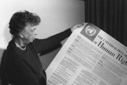 A person woman-presenting holds a large piece of paper covered with text and a large title reading "The Universal Declaration of Human Rights."