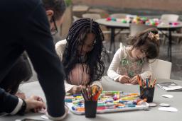 Two children sit at a table making art while an adult leans on the table, watching.