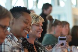 A group of people sit at a long dining table, laughing and enjoying a meal together. The focus is on two people smiling brightly - one wearing a plaid shirt and the other in a black patterned top, with glasses and plates arranged neatly in front of them.