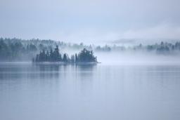 Mist rises off a large, calm lake surrounded by an evergreen forest.