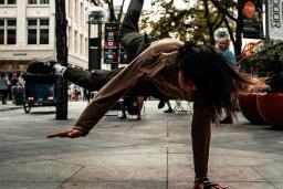 A woman in a grey jacket and darker grey pants is breakdancing on a sidewalk during daytime. Trees and buildings are in the background.