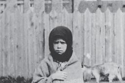 A young girl stands facing the camera with a sorrowful expression, clutching a shawl around her.