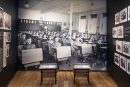 A museum exhibit that includes small school desks in front of a large photographic background of Indigenous children sitting at their desks in a classroom.