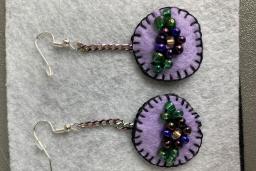 A pair of light purple felt earrings with dark purple sewn borders. In the middle of the earrings are beaded flowers.