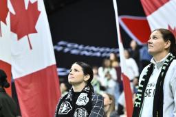 People stand on a stadium field during a pre-game ceremony as large Canadian flags are held behind them, filling the background with red and white.