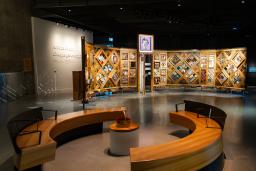 A museum exhibition featuring a large, curved freestanding wooden wall on which is mounted numerous small objects arranged in diamond and rectangular patterns. In the foreground, there circular benches creating an inviting space for visitors to view and contemplate the exhibit.