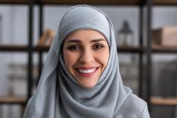 A woman wearing a blue hijab smiles toward the camera, with a bookshelf in the background.