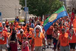 Hundreds of demonstrators in orange shirts, many also wearing traditional Indigenous articles such as a headdress or ribbon skirt and carrying signs, march down a wide paved street.