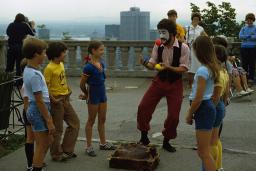 A man with a painted face juggles outdoors, as smiling children watch. A city skyline appears behind him at a distance.