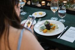 A woman with light skin and long, dark brown hair is using her cell phone to take a photo of a plate of gourmet food on a table.