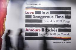 Three silhouettes of people walking past a large wall panel featuring the logo for the Love in a Dangerous Time exhibition. The silhouettes are blurred as the individuals are walking from left to right.