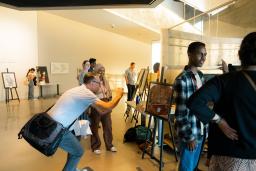 In a museum gallery, students exhibit projects on easels. Two students speak with adults about their work.