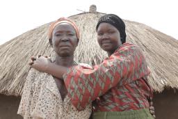 Two women are standing in front of a hut with a straw roof, looking at the camera. The younger woman on the right is smiling and embracing the one on the left.