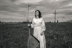 A woman in a long white dress stands in a field holding a bandura, a Ukrainian plucked string folk instrument.