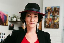A dark-haired, fair-skinned woman wears a black and scarlet hat, large earrings of white feathers, a bright red top and a black jacket with white prints on the sleeves. She has one hand on her hip and the other rests on a ruler on a design table.