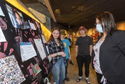 Three students stand in front of a poster board, placed on a table. A museum visitor stands in front of them looking at the poster board.