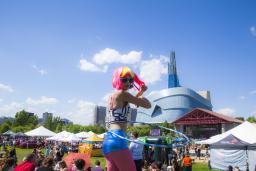 A person in brightly coloured wig and clothing swinging a hula hoop at an outdoor festival, with the Museum in the background.