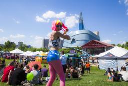A hula hoop dancer with a pink and fluorescent yellow wig with pink leggings walks in a crowd of people sitting on a field of grass in front of a large glass building.