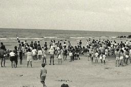 A black-and-white photograph of a crowd of people, most of them standing, on a beach.