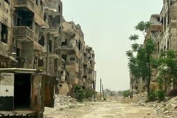 An abandoned city street where the building windows are gone, rubble lies piled in the road and a truck sits abandoned with its back doors open.